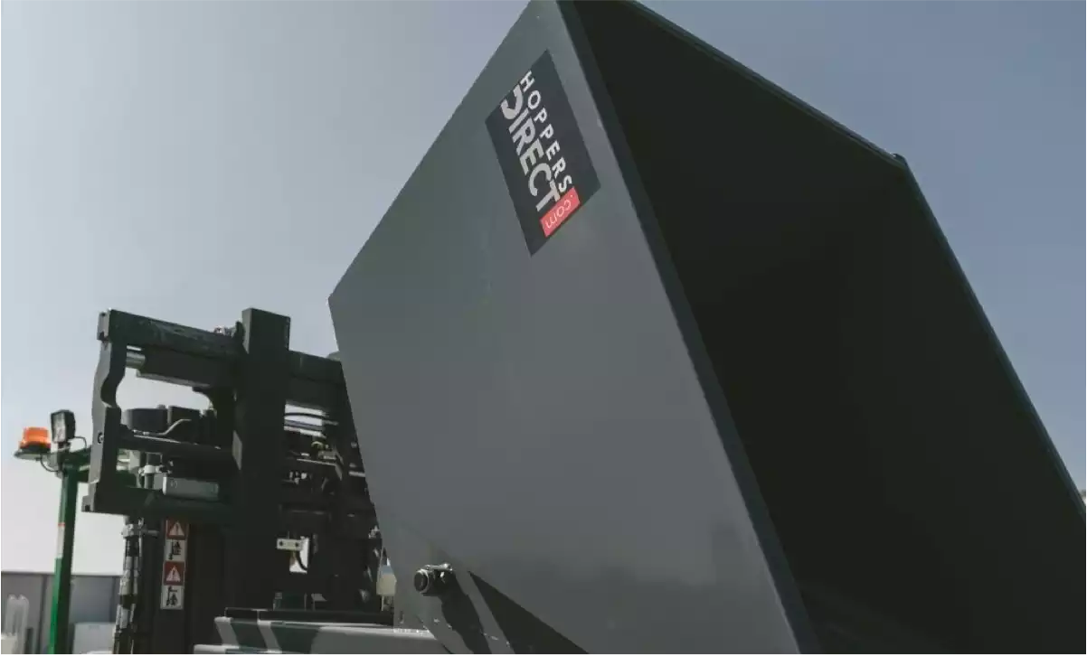 Close-up view of a black construction hopper attached to a forklift, highlighting the branding of Hoppers Direct. The image emphasizes the sturdy construction and design features of the self-dumping hopper against a clear blue sky.